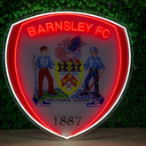 A large illuminated Barnsley FC shield sign mounted against a green leafy wall. The sign features a glowing red and white border, the club name in red neon at the top, a detailed crest in the center, and the year 1887 at the bottom.
