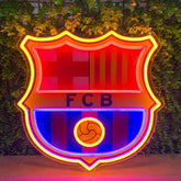 A large FC Barcelona neon sign glowing brightly against a background of green ivy-covered walls. The illuminated crest features red and blue stripes, yellow accents, and the letters 'FCB' in the center.