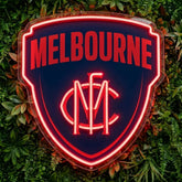 A shield-shaped Melbourne sports sign with glowing red neon lettering and logo on a dark navy background, mounted against a backdrop of green foliage.