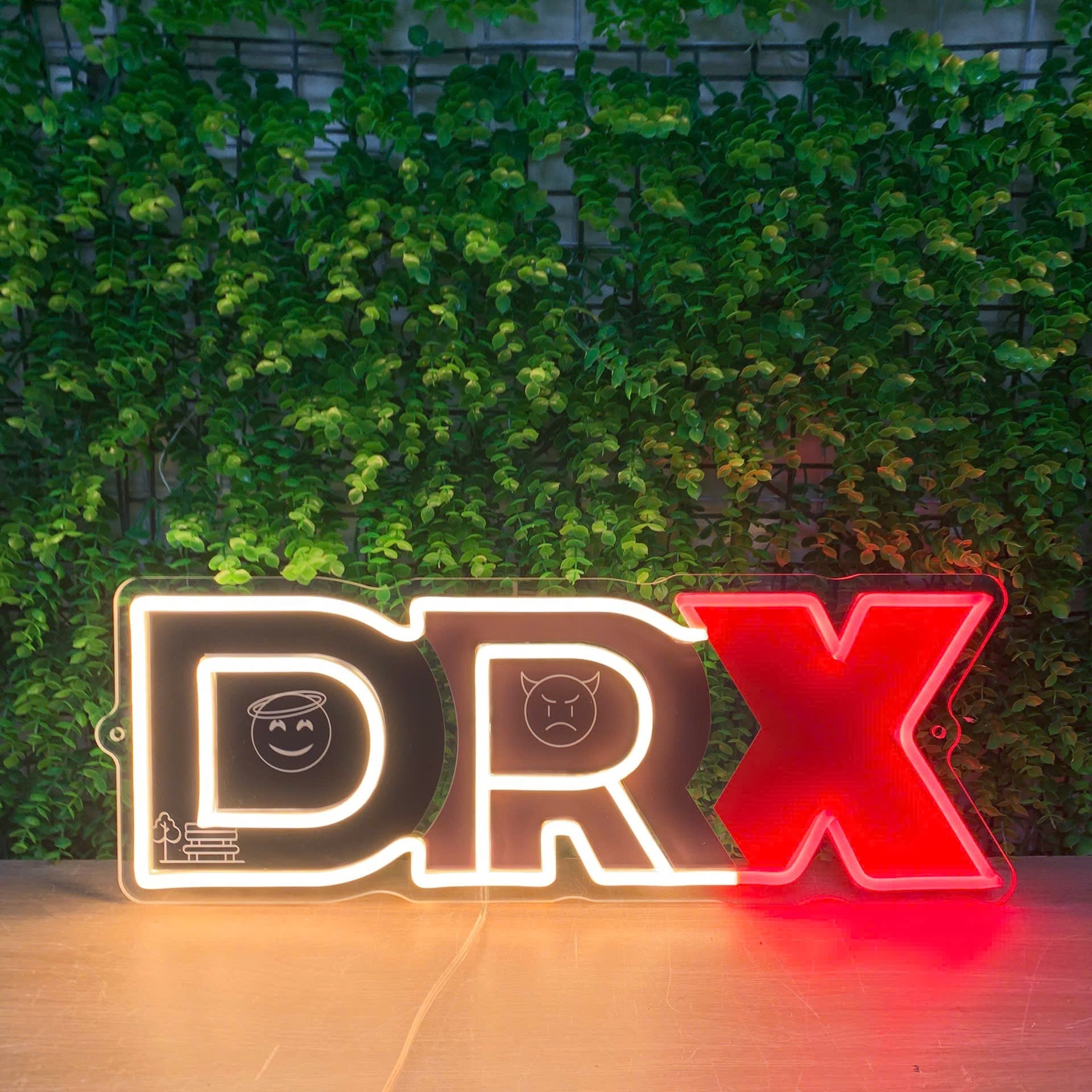 A neon-style decorative sign spelling "DRX" displayed on a tabletop in front of a green leafy wall. The letters are outlined in glowing light, with the first two letters in black and the final letter in bright red.