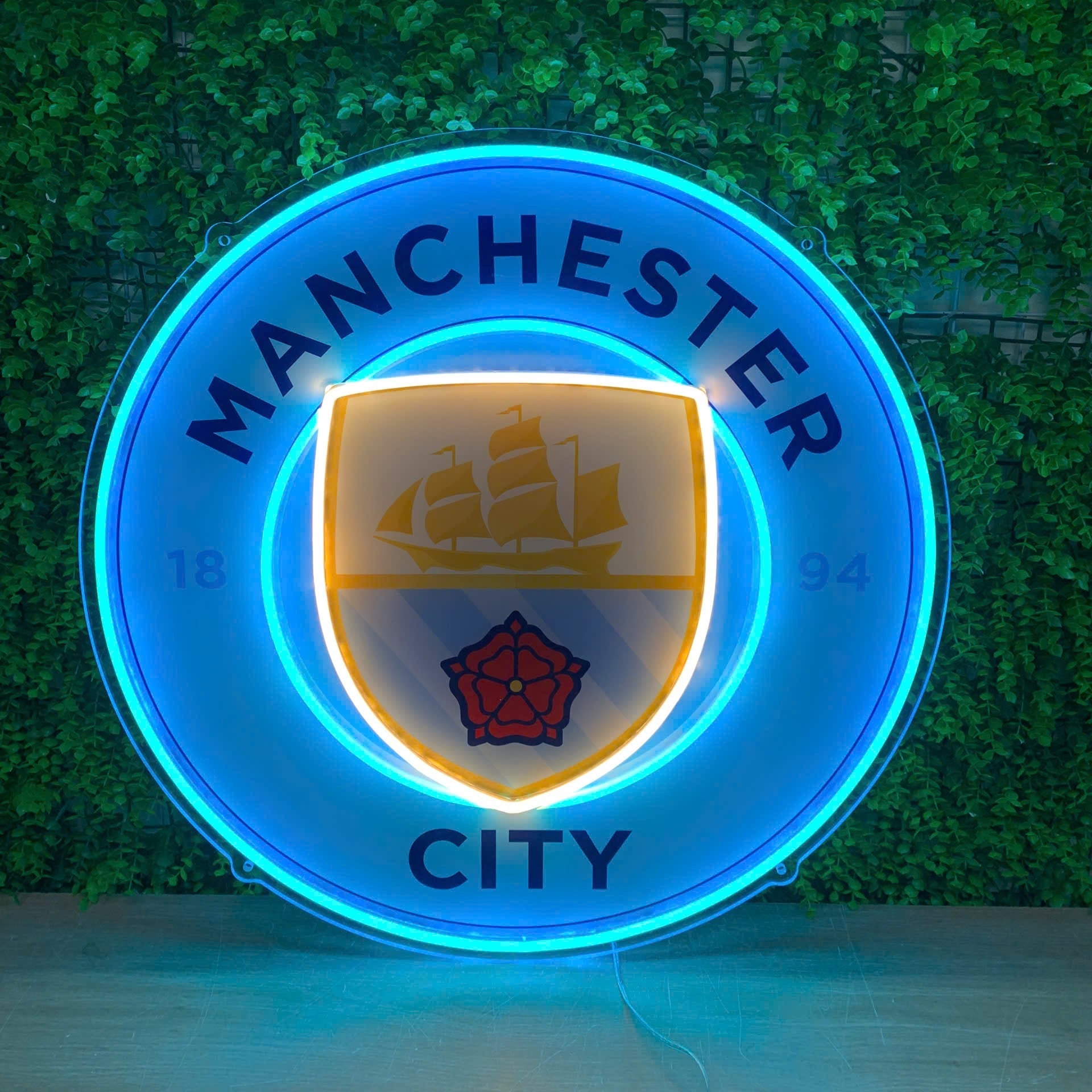 A glowing Manchester City neon-style sign mounted against a green ivy-covered wall. The circular sign features bright blue neon borders with the words MANCHESTER CITY, a central crest with a ship and red rose, and warm yellow light around the inner shield.