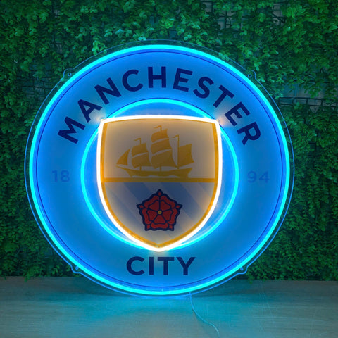 A glowing Manchester City neon-style sign mounted against a green ivy-covered wall. The circular sign features bright blue neon borders with the words MANCHESTER CITY, a central crest with a ship and red rose, and warm yellow light around the inner shield.