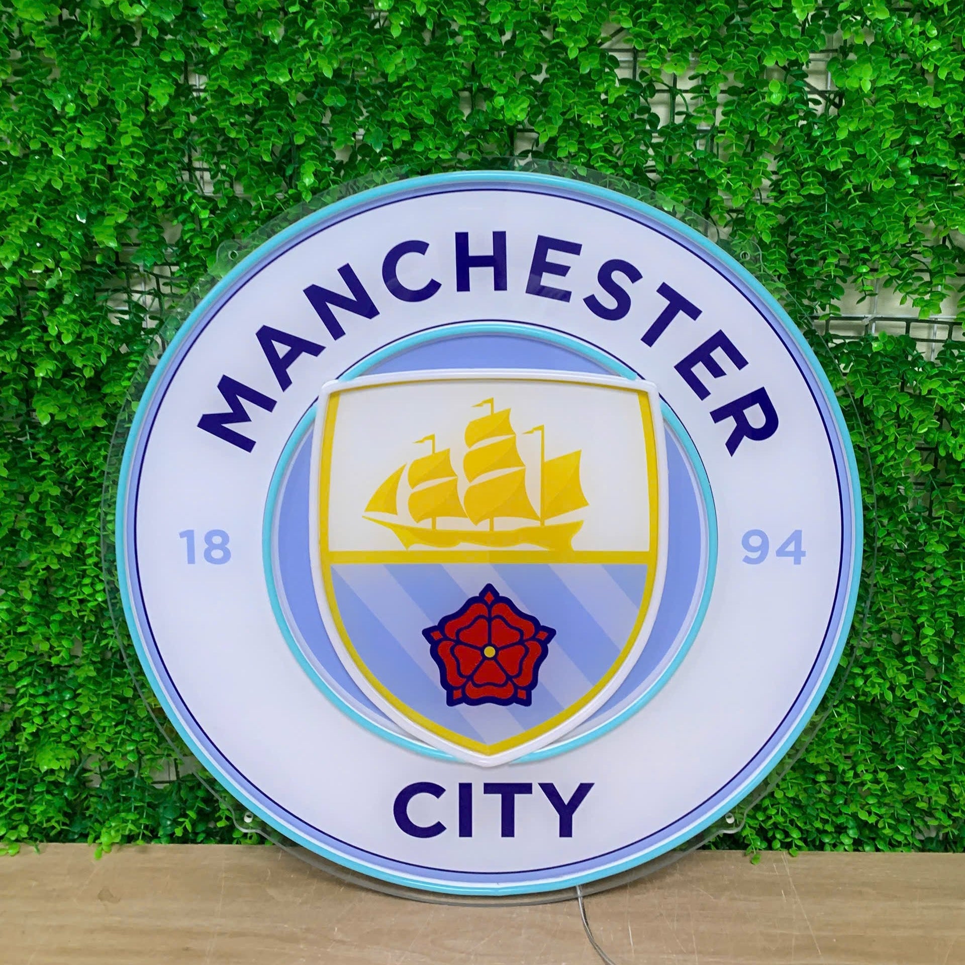 A circular Manchester City FC sign featuring the club's crest, displayed against a green artificial foliage wall.