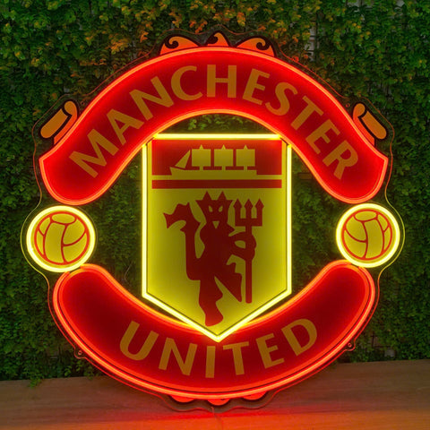A glowing Manchester United neon sign displaying the club's crest with illuminated red, yellow, and orange lights, set against a green ivy-covered wall.