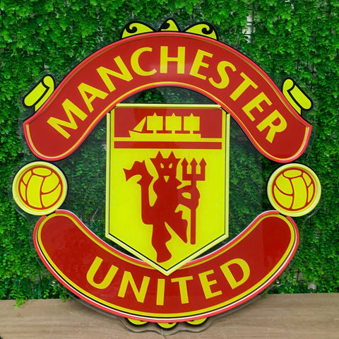 A large Manchester United crest wall sign with red and yellow colors, displayed against a green ivy wall backdrop.