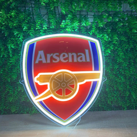 A glowing Arsenal football club shield sign with a red, blue, and gold cannon emblem, set against a green leaf wall background.